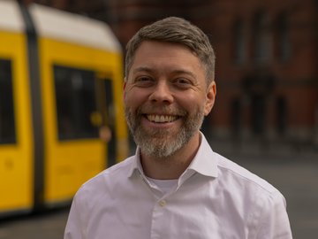 Auf dem Bild ist der Linke Bürgermeisterkandidat für Pankow, Jonas Teune, zu sehen. Er posiert im weißen Hemd. Im Hintergrund ist noch leicht das Pankower Rathaus zu erkennen. Außerdem ist dort eine gelbe Straßenbahn zu sehen.