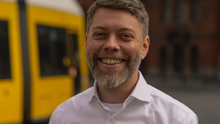 Auf dem Bild ist der Linke Bürgermeisterkandidat für Pankow, Jonas Teune, zu sehen. Er posiert im weißen Hemd. Im Hintergrund ist noch leicht das Pankower Rathaus zu erkennen. Außerdem ist dort eine gelbe Straßenbahn zu sehen.