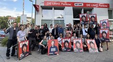 Vor dem Büro der LINKEN Pankow steht eine größere Gruppe von Menschen. Einige halten Wahlkampfplakate mit den Porträts von Elif Eralp und Jonas Teune in die Kamera. Auf den Die Gruppe wirkt gut gelaunt und motiviert; einige Personen heben die Faust. Im Hintergrund ist das Büro der LINKEN Pankow mit roter Beschriftung zu sehen. Die Aktion findet auf einem Gehweg vor dem Büro statt.