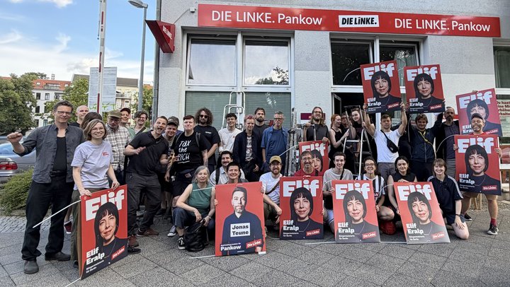 Vor dem Büro der LINKEN Pankow steht eine größere Gruppe von Menschen. Einige halten Wahlkampfplakate mit den Porträts von Elif Eralp und Jonas Teune in die Kamera. Auf den Die Gruppe wirkt gut gelaunt und motiviert; einige Personen heben die Faust. Im Hintergrund ist das Büro der LINKEN Pankow mit roter Beschriftung zu sehen. Die Aktion findet auf einem Gehweg vor dem Büro statt.