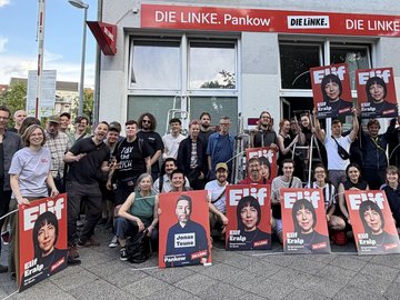 Vor dem Büro der LINKEN Pankow steht eine größere Gruppe von Menschen. Einige halten Wahlkampfplakate mit den Porträts von Elif Eralp und Jonas Teune in die Kamera. Auf den Die Gruppe wirkt gut gelaunt und motiviert; einige Personen heben die Faust. Im Hintergrund ist das Büro der LINKEN Pankow mit roter Beschriftung zu sehen. Die Aktion findet auf einem Gehweg vor dem Büro statt.