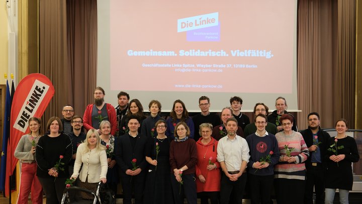Eine Gruppe von Menschen steht gemeinsam für ein Gruppenfoto im Pankower BVV-Saal. Es handelt die um die Linken Kandidat*innen für die anstehende Wahl zur Bezirksverordnetenversammlung. Viele halten einzelne rote Rosen in den Händen. Im Hintergrund ist auf einer Leinwand das Logo „Die Linke – Bezirksverband Pankow“ sowie der Slogan „Gemeinsam. Solidarisch. Vielfältig.“ zu sehen. Links im Bild steht ein rotes DIE-LINKE-Banner. Die Personen stehen in zwei Reihen und schauen überwiegend in die Kamera.