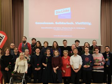 Eine Gruppe von Menschen steht gemeinsam für ein Gruppenfoto im Pankower BVV-Saal. Es handelt die um die Linken Kandidat*innen für die anstehende Wahl zur Bezirksverordnetenversammlung. Viele halten einzelne rote Rosen in den Händen. Im Hintergrund ist auf einer Leinwand das Logo „Die Linke – Bezirksverband Pankow“ sowie der Slogan „Gemeinsam. Solidarisch. Vielfältig.“ zu sehen. Links im Bild steht ein rotes DIE-LINKE-Banner. Die Personen stehen in zwei Reihen und schauen überwiegend in die Kamera.